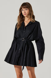 A woman is wearing a black, long-sleeved shirt dress made from a shiny, satin-like fabric. The dress features a pointed collar, button-down front, and a matching belt with a circular metal buckle at the waist. The skirt portion of the dress is gathered and flares out slightly, ending above the knees.